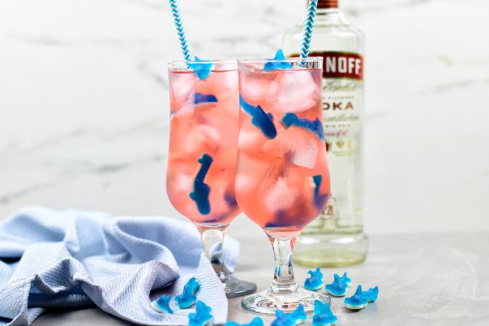 Two shark attack punch cocktails with bottle of vodka behind them and many shark gummies surrounding on the ground. 