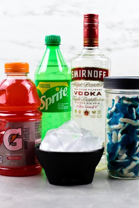 Ingredients shown: punch Gatorade, sprite soda bottle, Smirnoff vodka bottle, jar filled with shark gummies, and black bowl of ice. 
