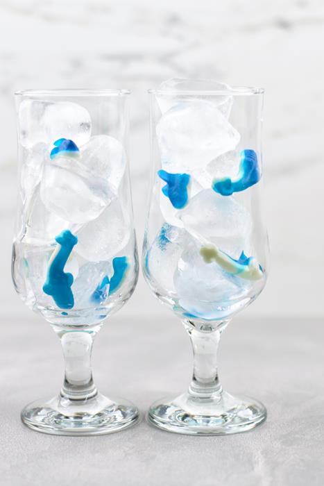 Two tall clear glasses with ice and shark gummies throughout it. 