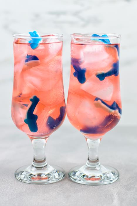 Two tall clear glasses with ice, gummies, and pink punch and sprite added. 