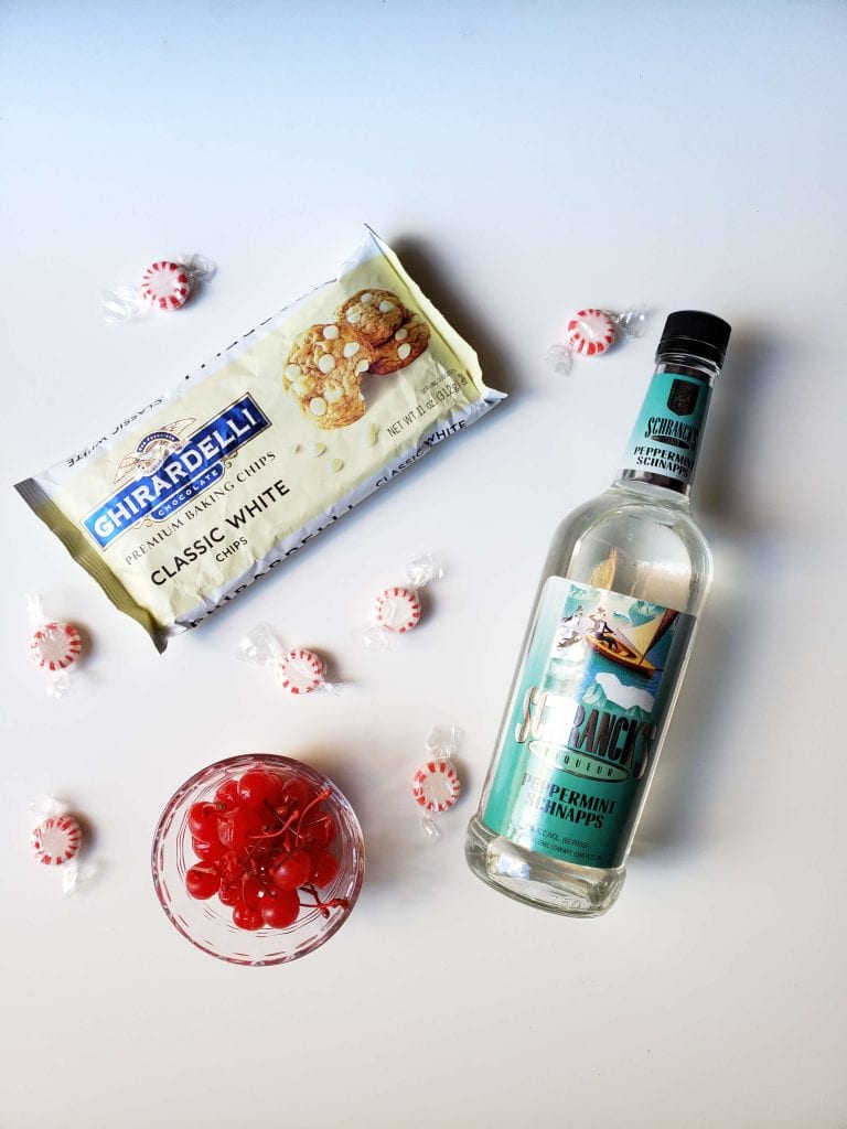 Above view of ingredients: clear bowl of maraschino cherries, bag of white chocolate chips, bottle of peppermint schnapps, and various peppermint candies wrapped up. 