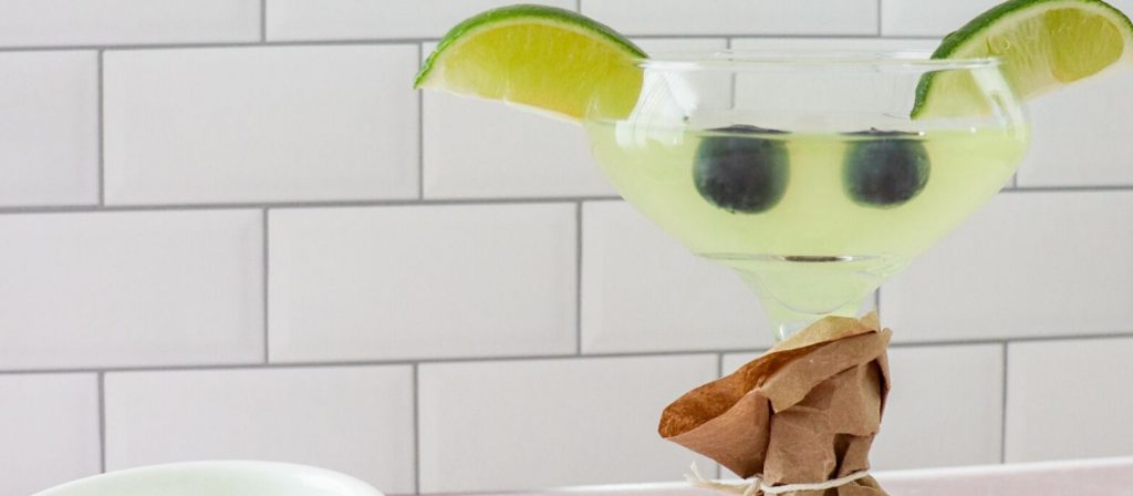 Close up of baby yoda cocktail showing green drink in a martini glass with two blueberries as eyes and limes on rims looking like ears. 