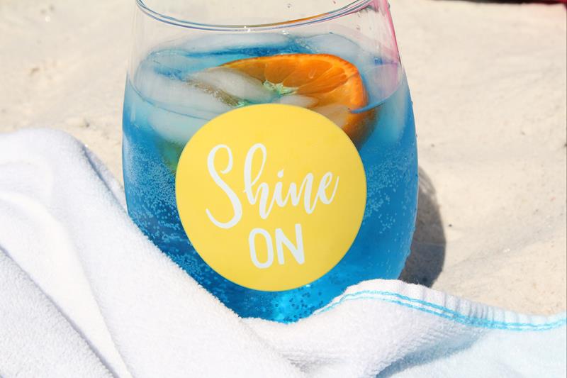close up of beach waves cocktail in a clear glass with yellow circle with white font that says shine on on a sandy beach/ 