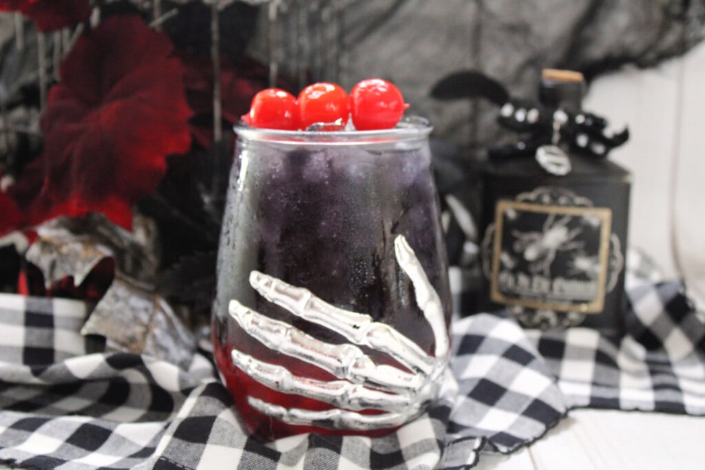Black Magic Cocktail shown in a clear glass with a skull hand on it. with three cherries as garnish. 