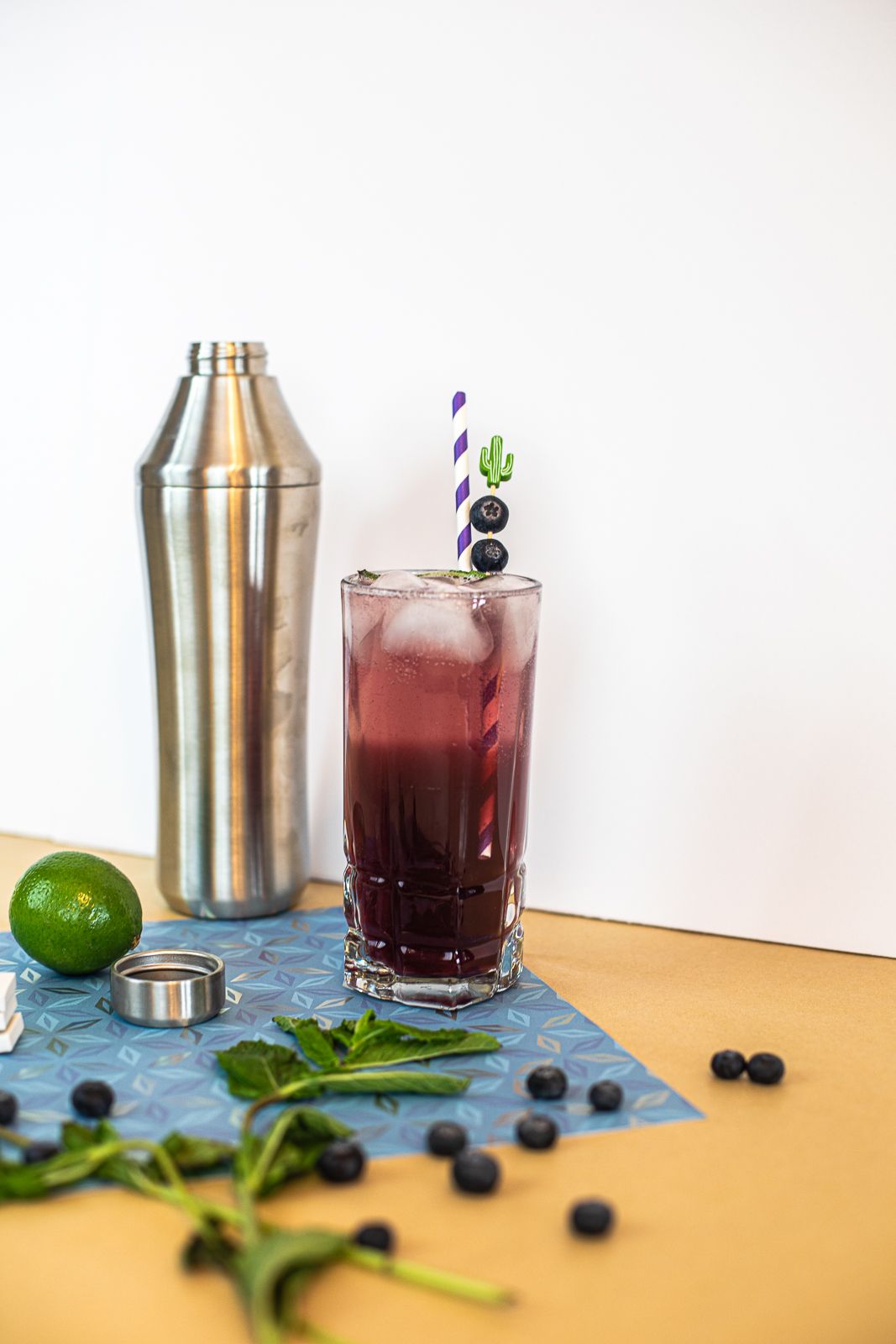 Shaker cup, cocktail, lime, mint, and blueberries shown. 