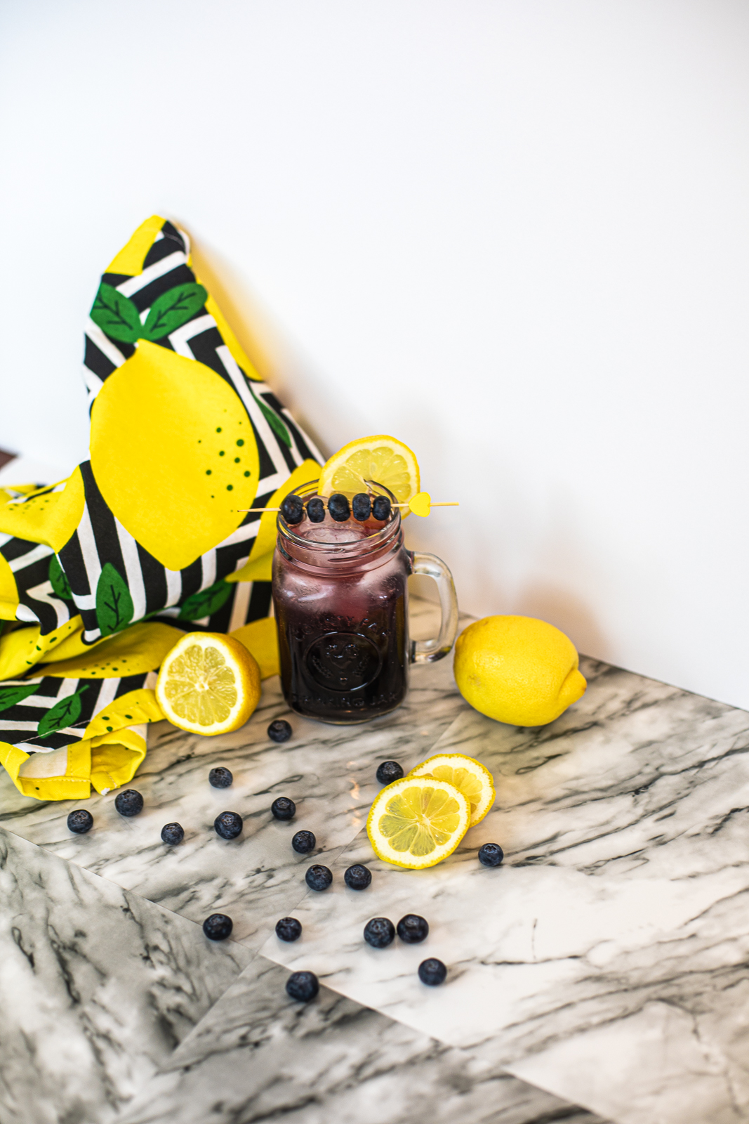 Finished cocktail on a marble floor with lemons and blueberries all around it. 