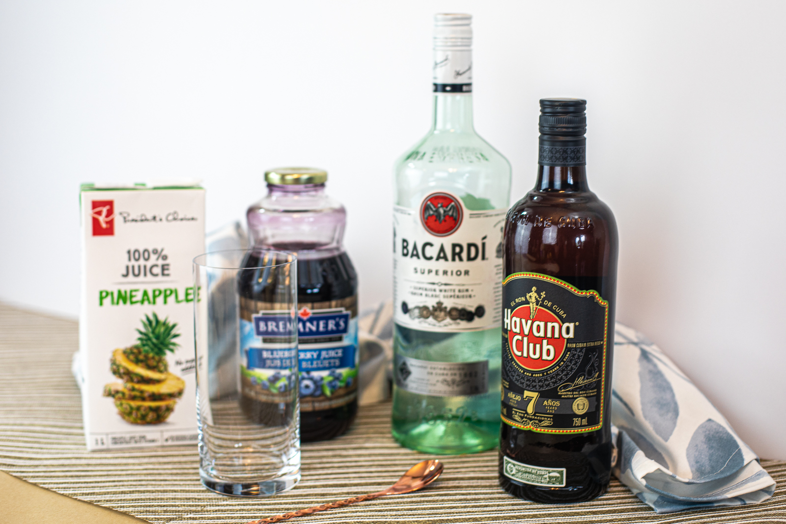 Ingredients required to make a Blueberry Rum Swizzle: pineapple juice, blueberry juice, glass, swizzle stick white rum, and amber rum. 