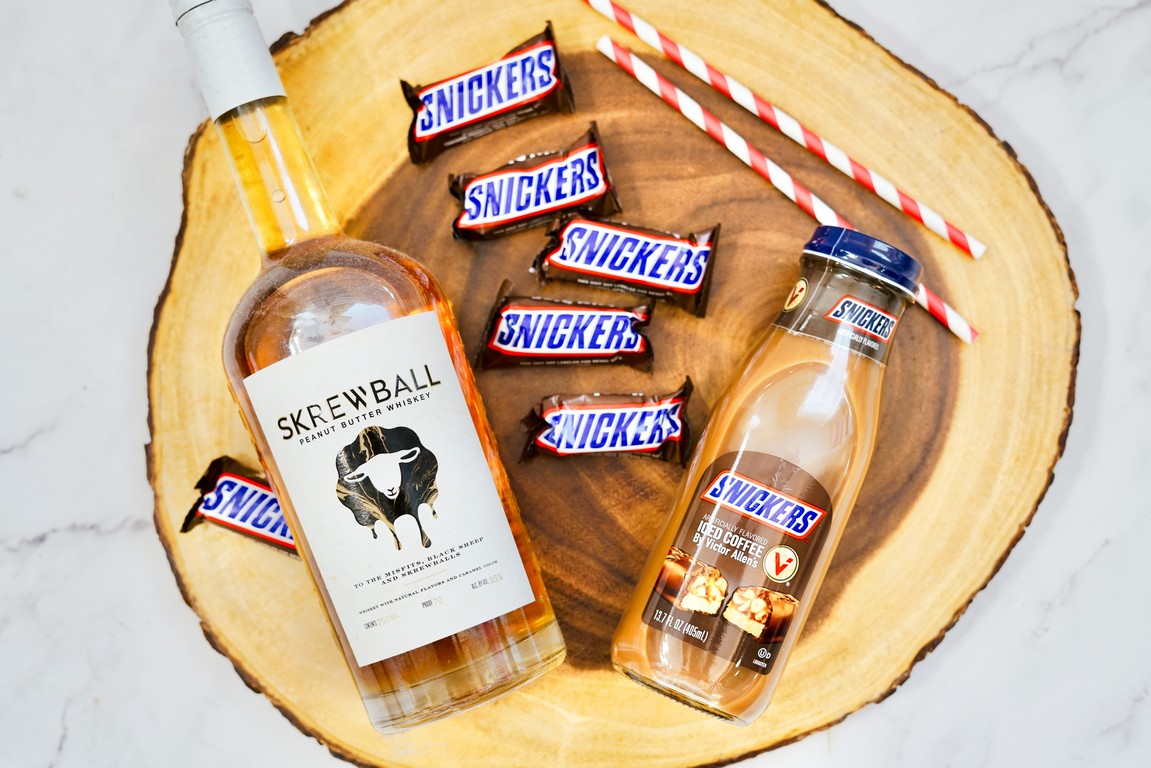 Ingredients needed for Boozy Snickers Iced Coffee on a wooden slab - peanut butter whiskey bottle, snickers iced coffee bottle, mini snickers chocolate bars, and two straws. 