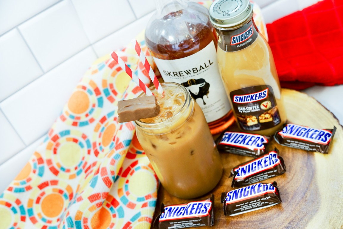 Boozy Snickers Iced Coffee - on a wooden round slab with mini chocolate bars, iced coffee, and whiskey bottles around it. 