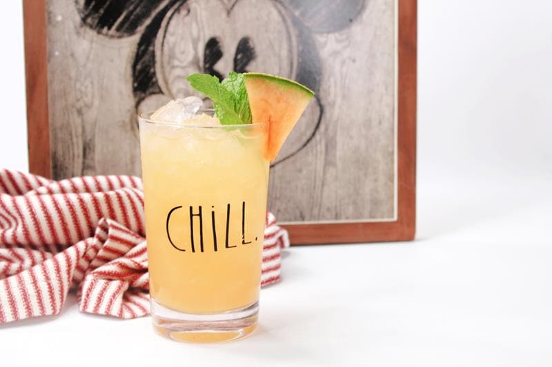 Disney Copycat Cantaloopy Cocktail in a tall clear glass that has black writing that says CHILL on it. With Cantaloupe and mint leaves as garnish. 