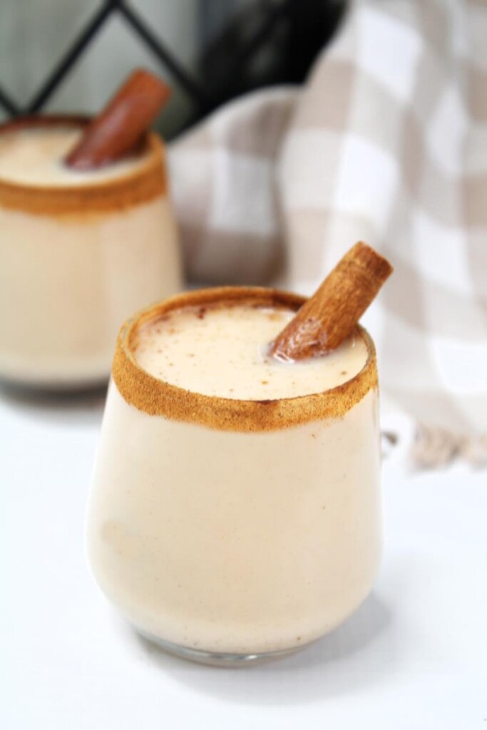 Slightly above shot of Churro shot with cinnamon around rim of clear glass filled with creamy drink with a cinnamon stick sticking out. 