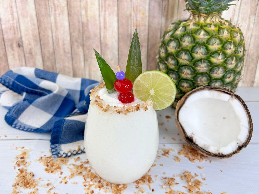 Close up of cocktail with a lime wheel, two cherries, and pineapple stem with a  coconut rim. 