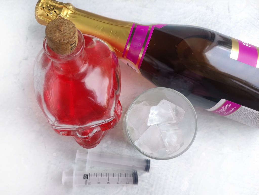 Ingredients needed - a skull glass with grenadine in it, glasses with ice in it, white sparkling wine bottle, and disposable syringes. 