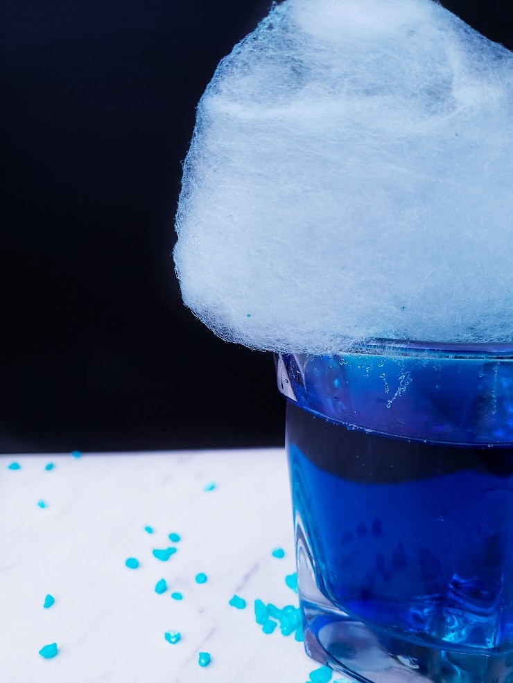 Close up of half a cocktail glass with blue drink and light blue cotton candy on top. 