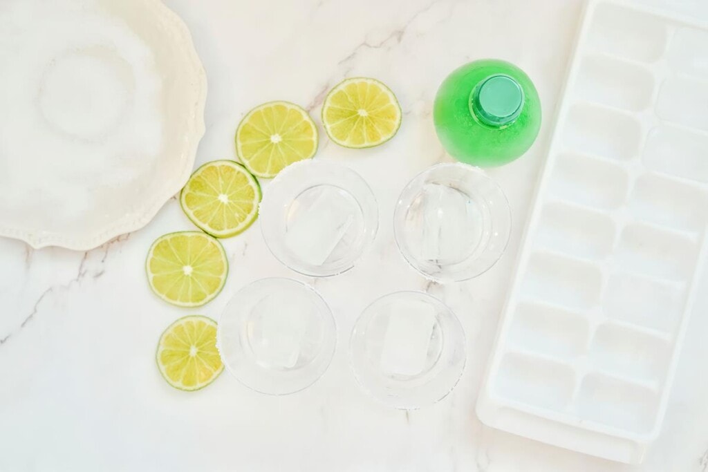 Above view of four salted rimmed clear glasses with ice added with now empty ice cube tray and sliced limes beside them. 
