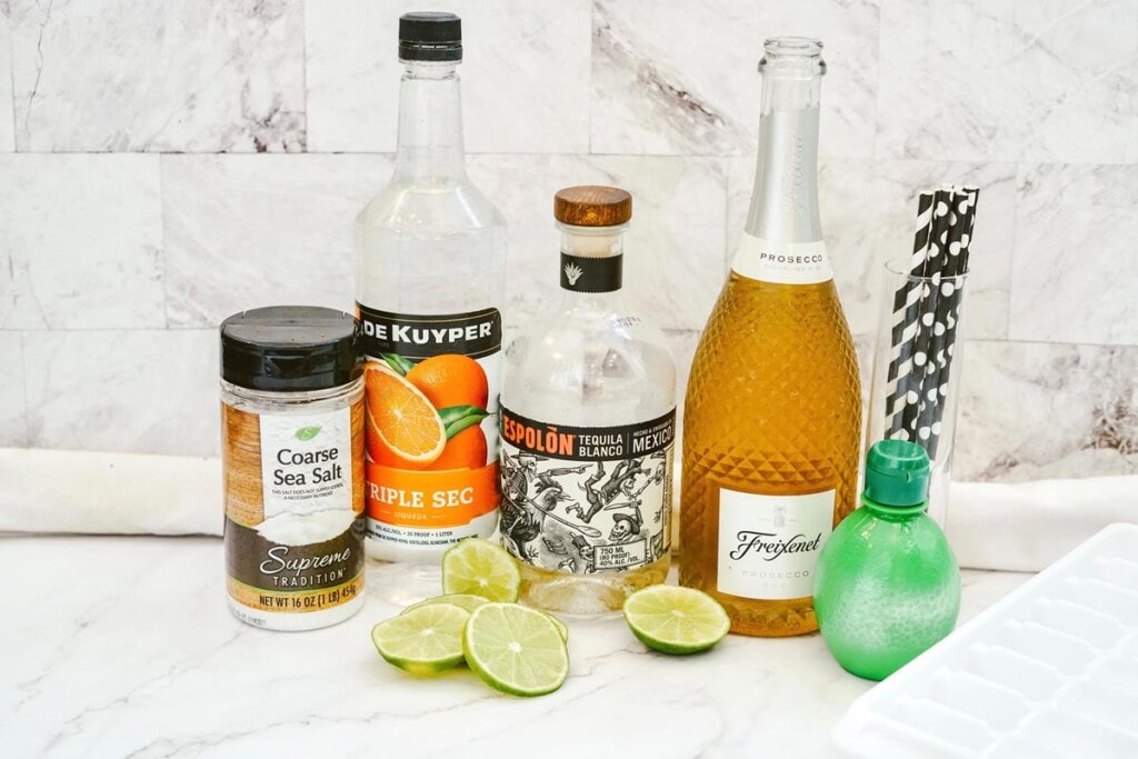 Photo showing ingredients for margarita: Bottle sea salt, bottle of triple sec, tequila bottle, prosecco bottle, tall glass with black and white straws, lime juice bottle, and sliced limes in front with ice cube tray beside. 
