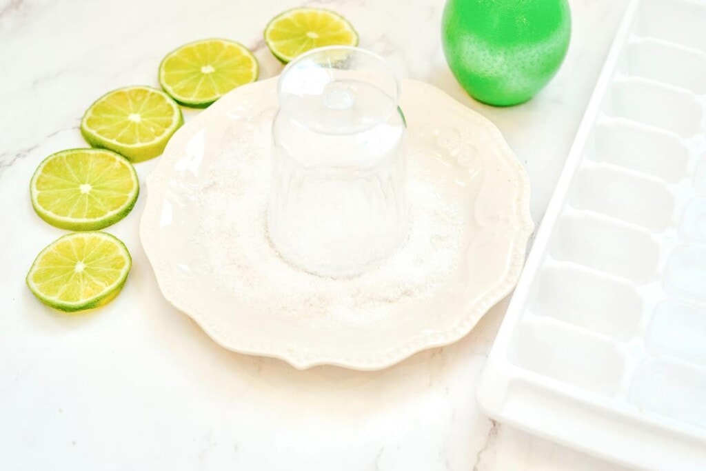 White plate with salt on it with upside down glasses having salt added to rim with ice cube tray beside it and cut up limes. 