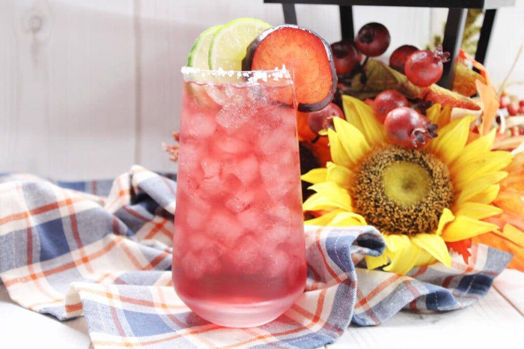 Showing plum margarita on a checkered red, white, and blue kitchen towel with sunflowers both yellow andorange with fake grapes in the background. 