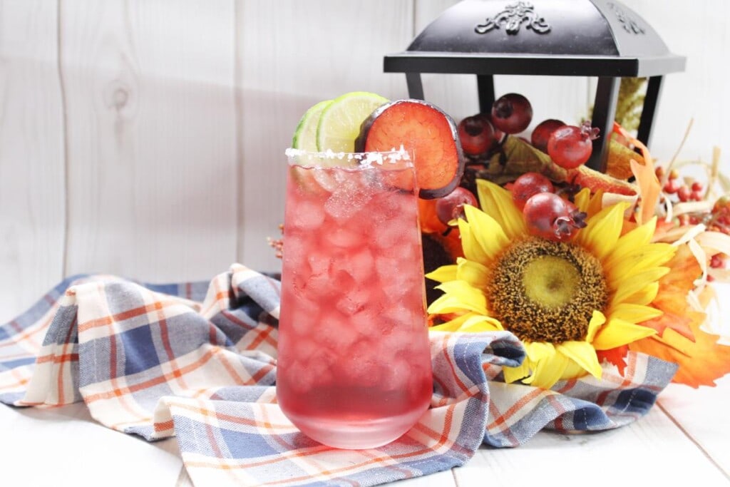 Plum margarita on a table with various fake flowers, fruit, and lantern in the background. 