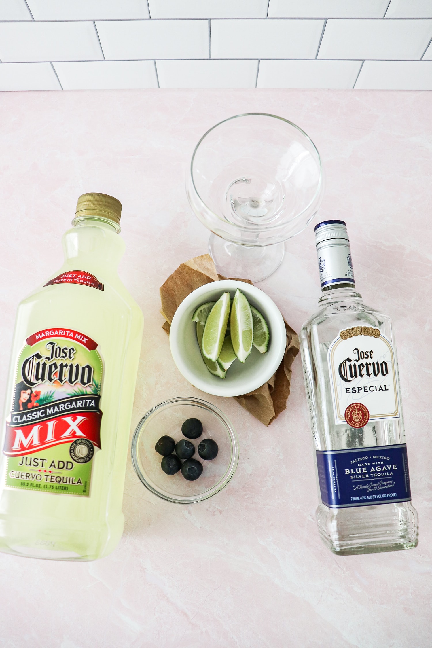 Above view of ingredients required for Baby yoda cocktail. bottle of classic margarita mix, white bowl of lime slices, clear bowl of blueberries, and bottle of tequilla. 