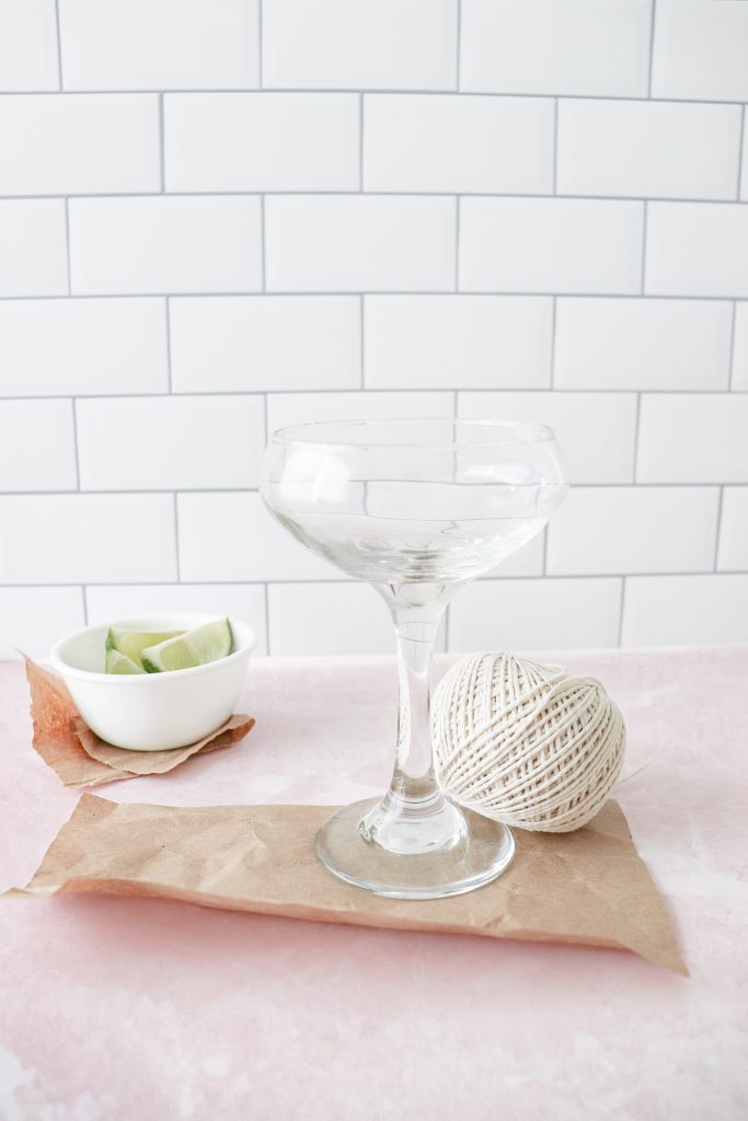 Brown paper under a clear tall martini glass with yarn and white bowl of lime slices beside. 