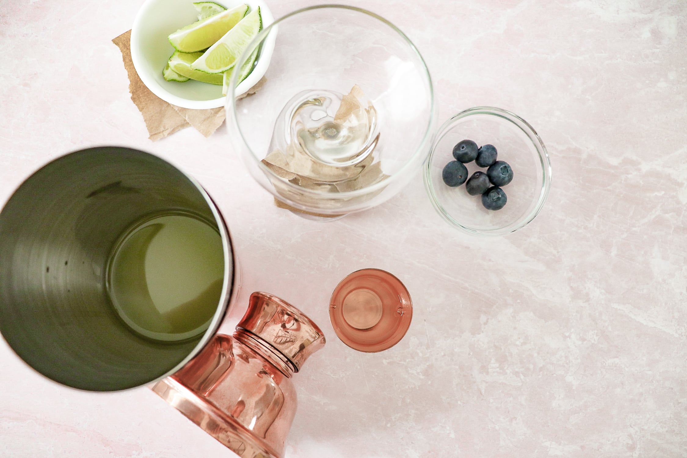 Above view of martini glass wrapped in brown paper, with white bowl of lime slices, white bowl of blueberries, and shaker cup with drink in it. 