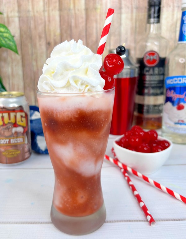 Root beer rum slush - redish brown slushy drink in tall clear glass topped with whipped cream and cherries. 