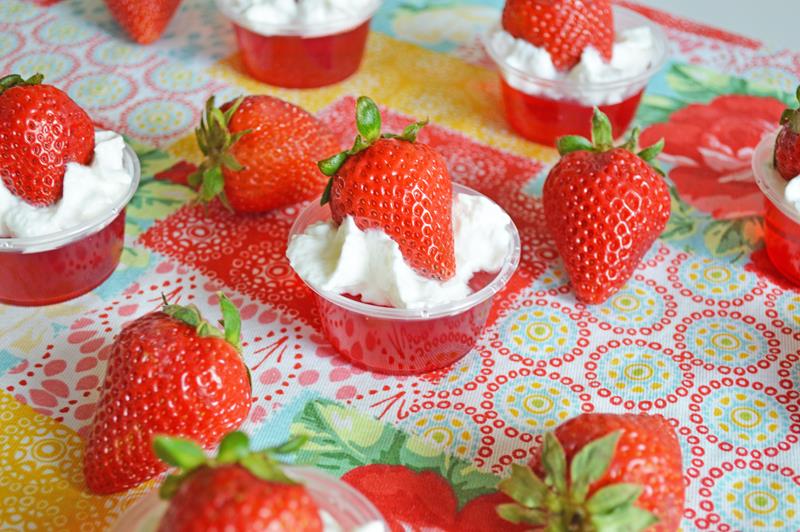 Strawberry Jello Shots on a table topped with whipped cream and a full strawberry surrounded by many fill strawberries 