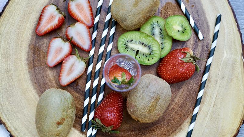 above view of shooter glass with strawberry in it, black and white straws, cut up strawberries and kiwi surrounding shooter glass 