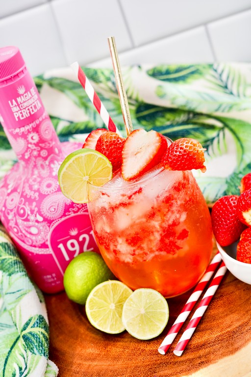Close up of soda with pink tequila bottle behind it with limes, strawberries and straws on table around it. 