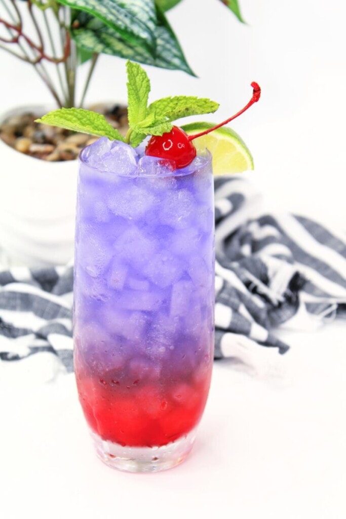 close up on cocktail in long clear glass with ice, pruple and red drink with mint, cherries, and lime as garnish