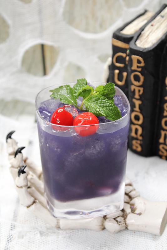 Slightly above view of Black Pearl cocktail with fresh mint and cherries as garnish on a skull hand coaster. 
