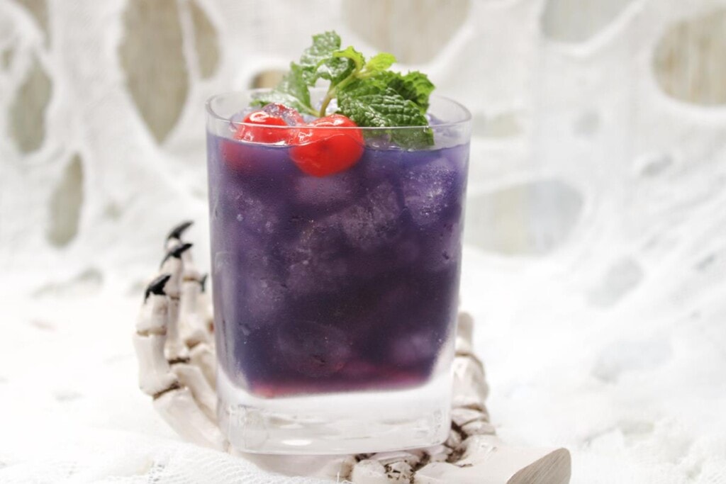 The Black Pearl Cocktail Recipe: Close up of dark purple cocktail filled with lots of ice with fresh mint and cherries as garnish. 