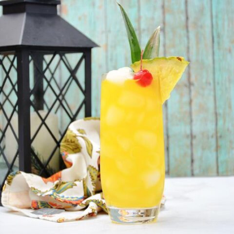 tropical whisky sour with cherries, ice, and pineapple wedge in front of an outdoor candle lamp