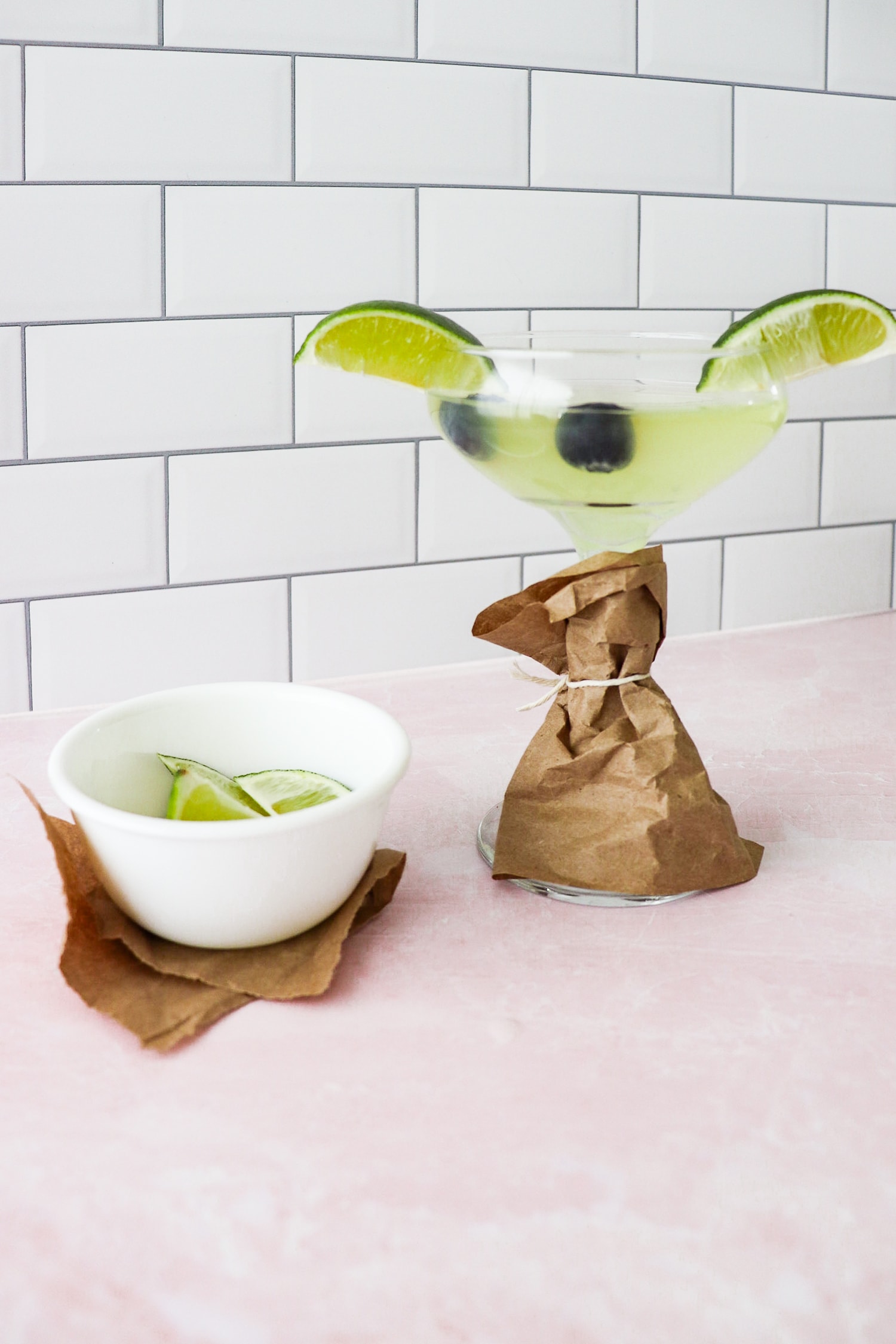 Martini glass wrapped with brown paper with blueberries in drink as eyes and limes as ears. 