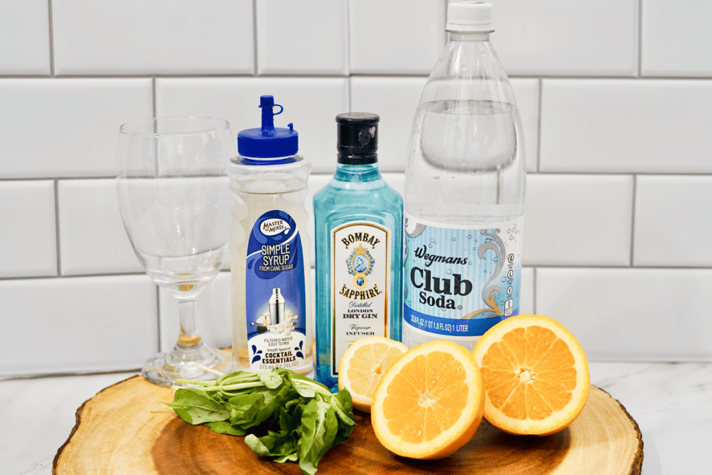 Wooden slab with all the ingredients need to completed your Orange Gin Basil Smash - glass, simple syrup, gin, club soda, oranges, and basil leaves. 