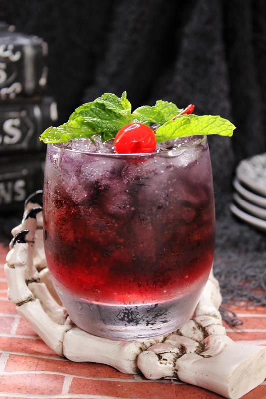 Showing red bottom layer of cocktail and dark blackish purple top layer filled with ice and topped with mint leaves and a cherry. 