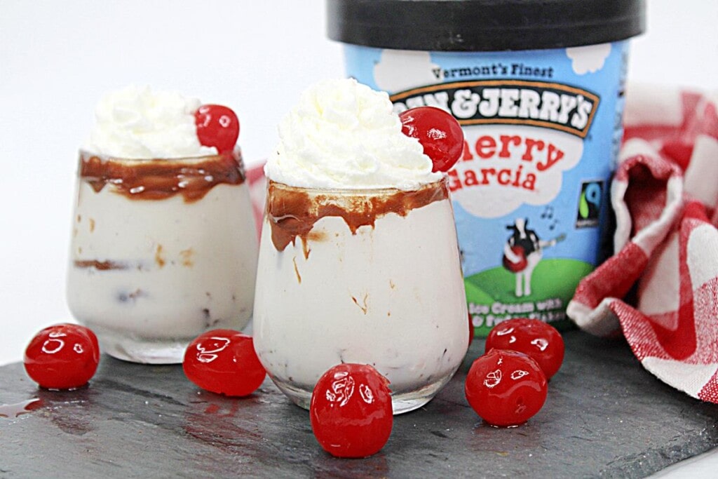 Cherry Garcia Ice Cream Shooters: Two completed shooters in stemless wine glasses, showing tan creamy drink with chocolate sauce around the rim topped with whipped cream and a cherry. 