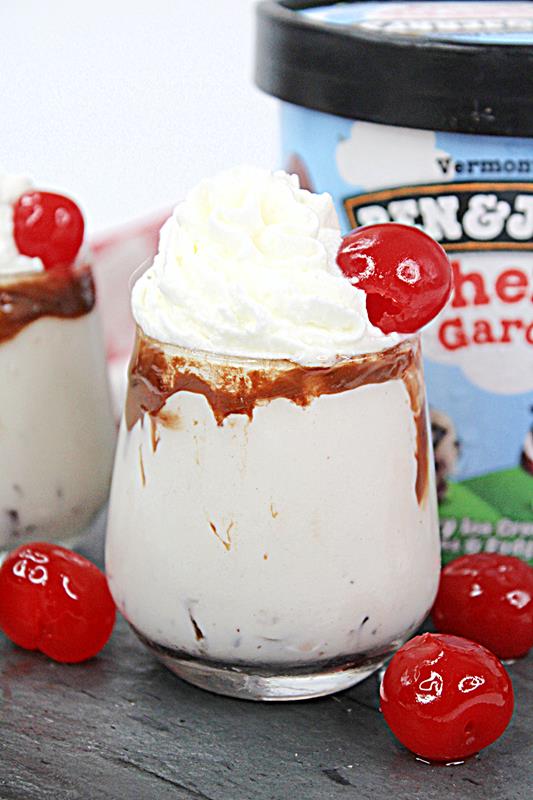 Creamy shooter with chocolate sauce on the rim with whipped cream on top and one cherry, with three other cherries on the ground around it. 