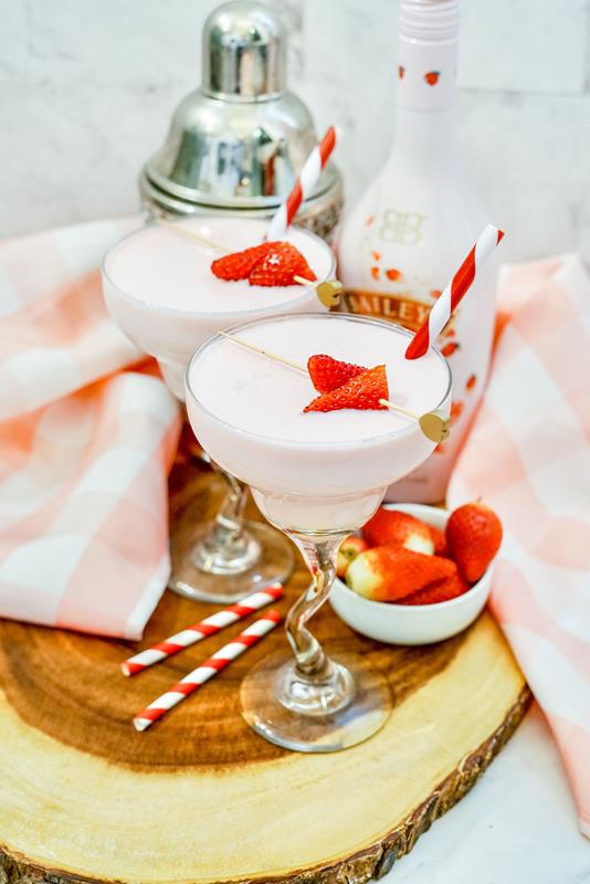 Light pink drink in a clear martini glass with two pieces of strawberries on top with a red and white straw sticking out. 