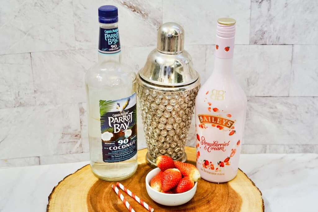 Ingredients on a wooden round board, white bowl of full strawberries, bottle of baileys strawberries and cream, fancy shaker cup, coconut rum, and two red and white straws. 