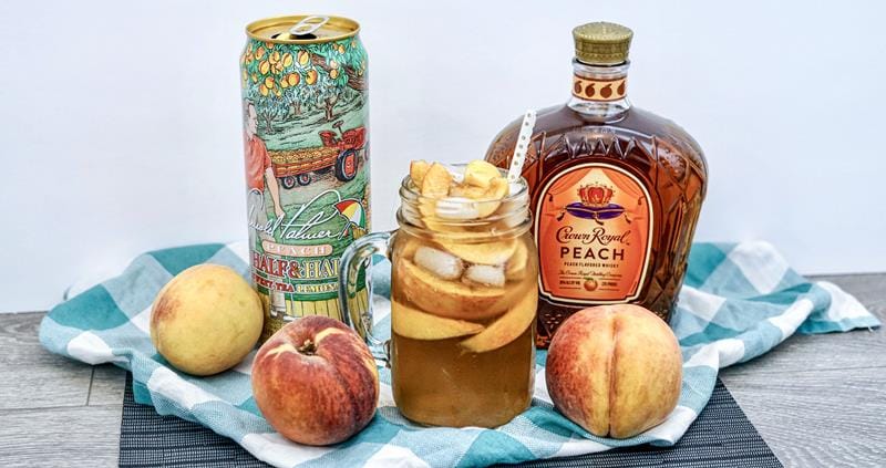 Crown peach tea drink in clear glass with ice and peach slices surrounded by full peaches, tea can, and crown royal bottle