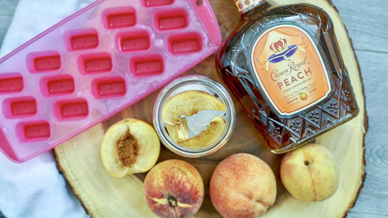 showing above shot of finished drink surrounded by empty pink ice cube tray, 3 peaches, and peach crown royal bottle 