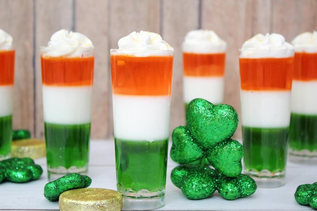 Irish Flag Jello Shots Recipe - Six Irish Flag Jello Shots, Tall glass shot glasses, green bottom layer, middle white layer, orange top layer. All topped with Whipped Cream. 