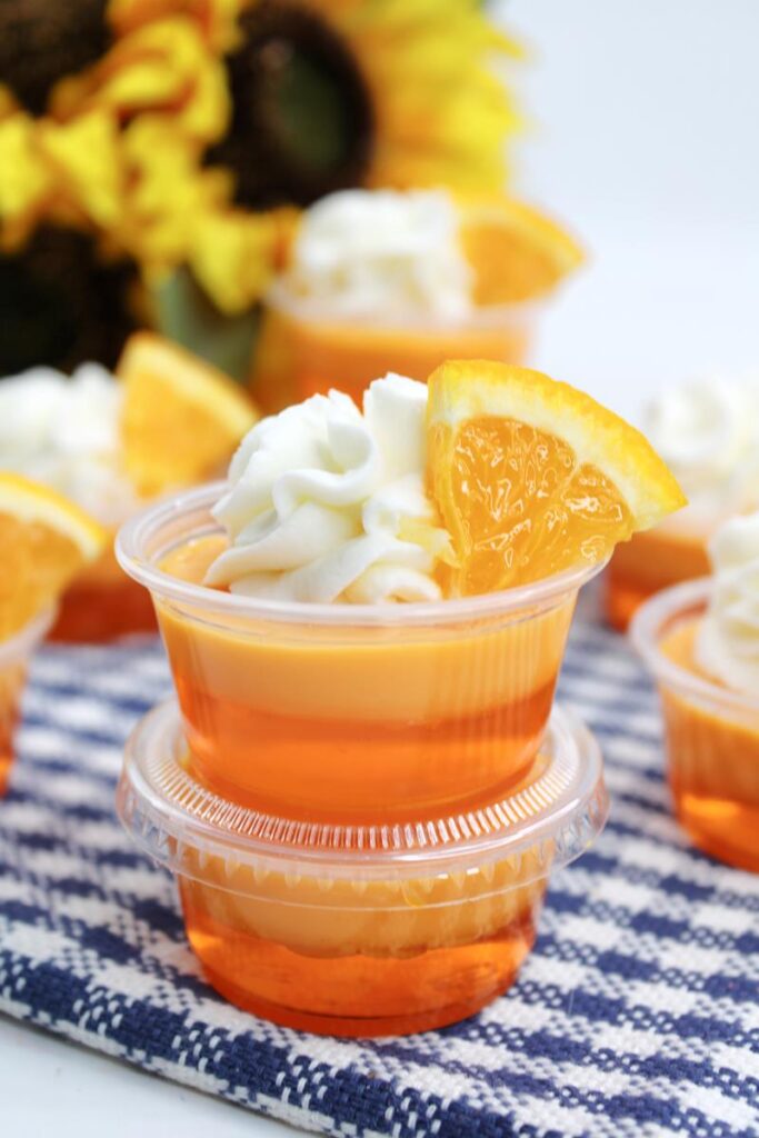 close up of two Orange Mango Daiquiri Jello Shots showing whipped cream and piece of orange as garnish. 