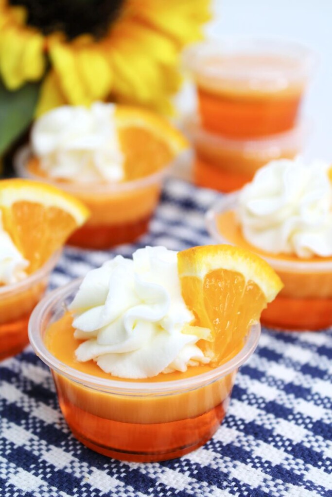 close up of jello shot showing orange clear bottom layer, solid orange second layer and whipped cream and orange slice as garnish. 