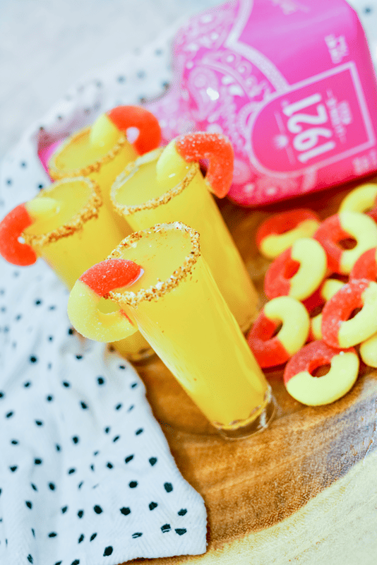 Peach Margarita Shooters - Four shooters with peach rings all around them and a bottle of Tequila in the background. 