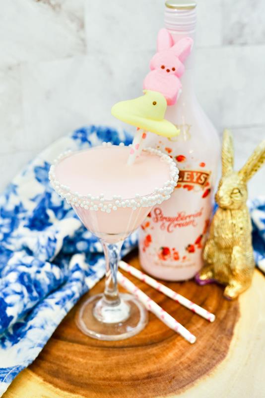 Completed Peeps martini with white round sprinkles on the rum and a yellow peep and pink bunny peep on a straw as garnish with strawberries and cream baileys bottle in the background and a gold wrapped chocolate bunny. 