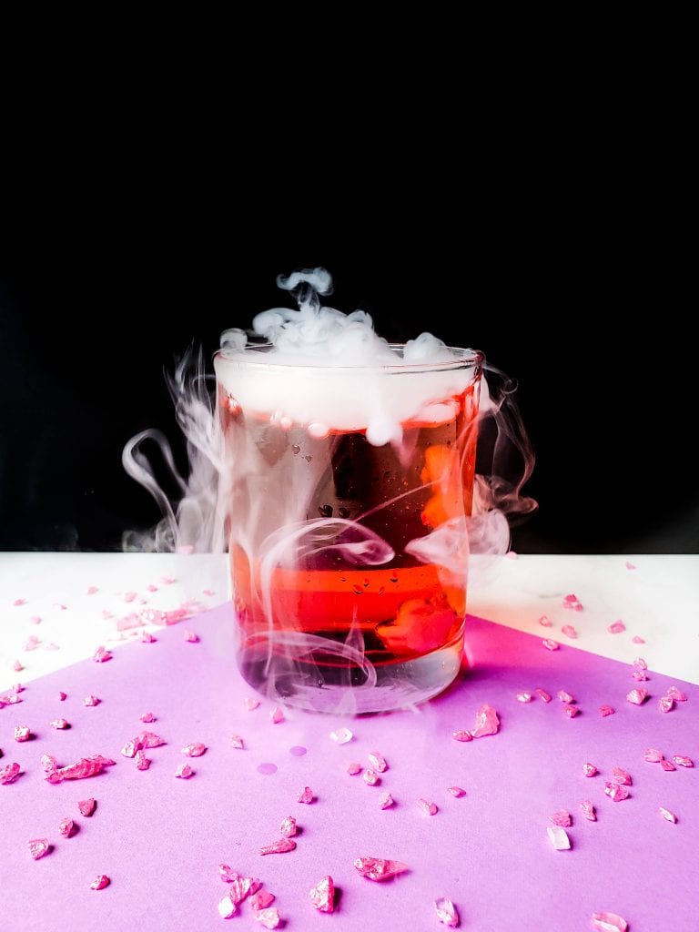 Cocktail in clear glass showing pinkish liquid with mist flowing out from it. 