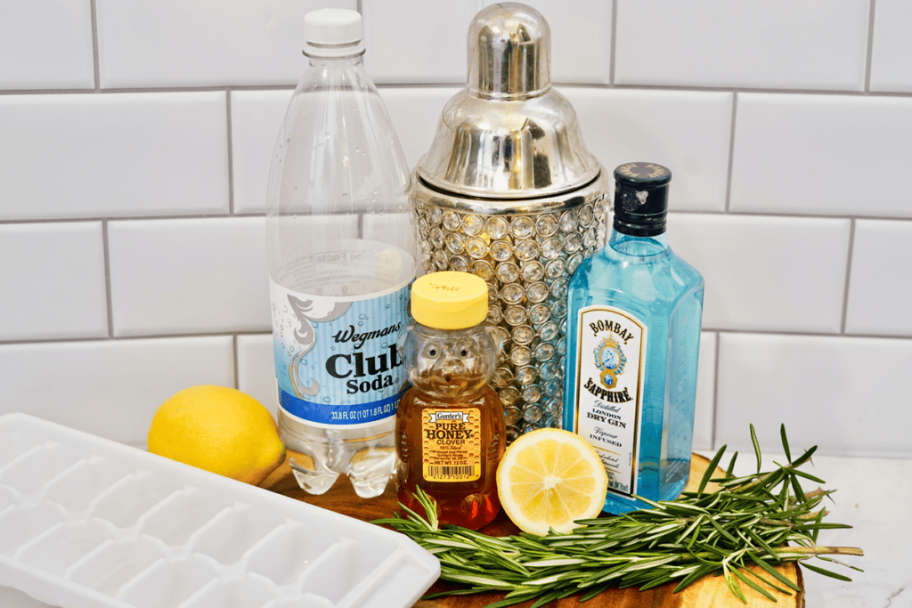 Rosemary Gin Fizz - Ingredients needed for cocktail, club soda, honey, shaker, gin, ice cube trays, lemons, rosemary all on a round wooden slab. 